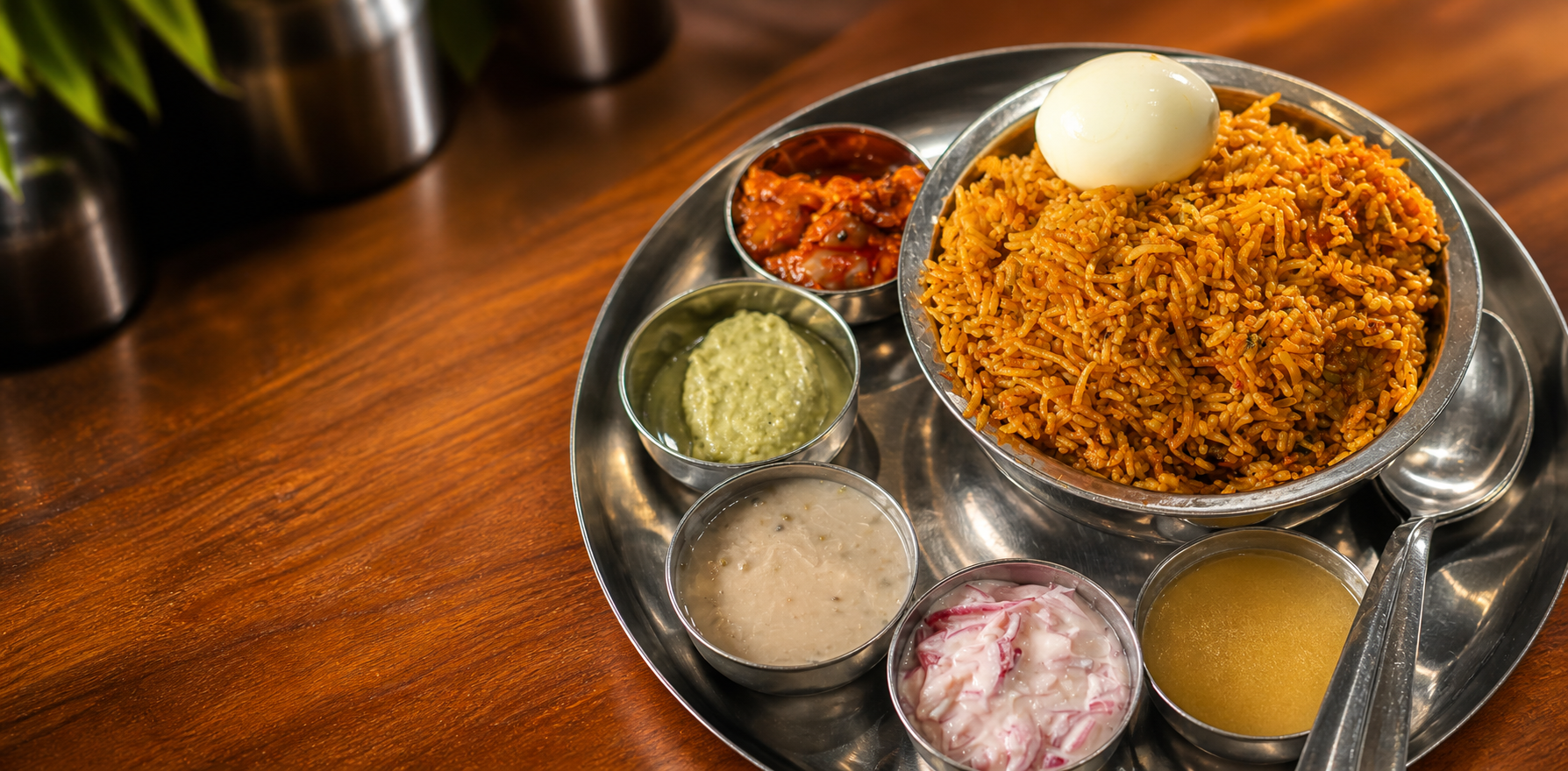 Briyani rice topped with a boiled egg, served with chutney, curry and sliced onions on a steel tray.