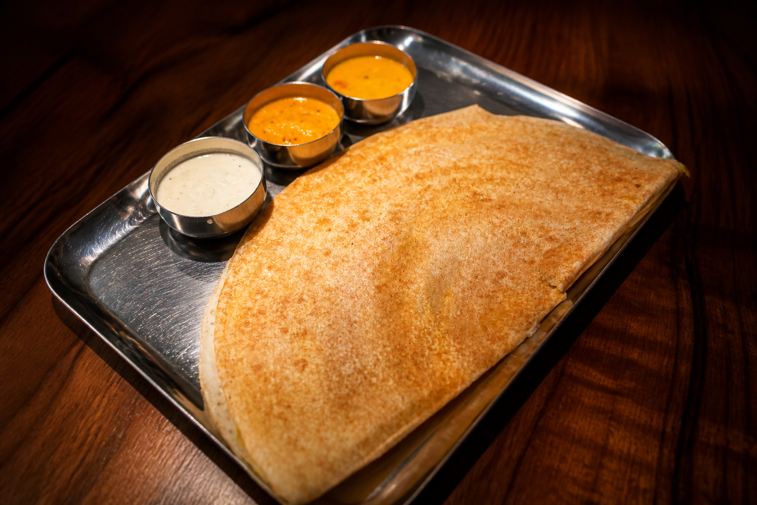 Large crisp dosa folded across a steel tray with two small bowls of chutney and curry.